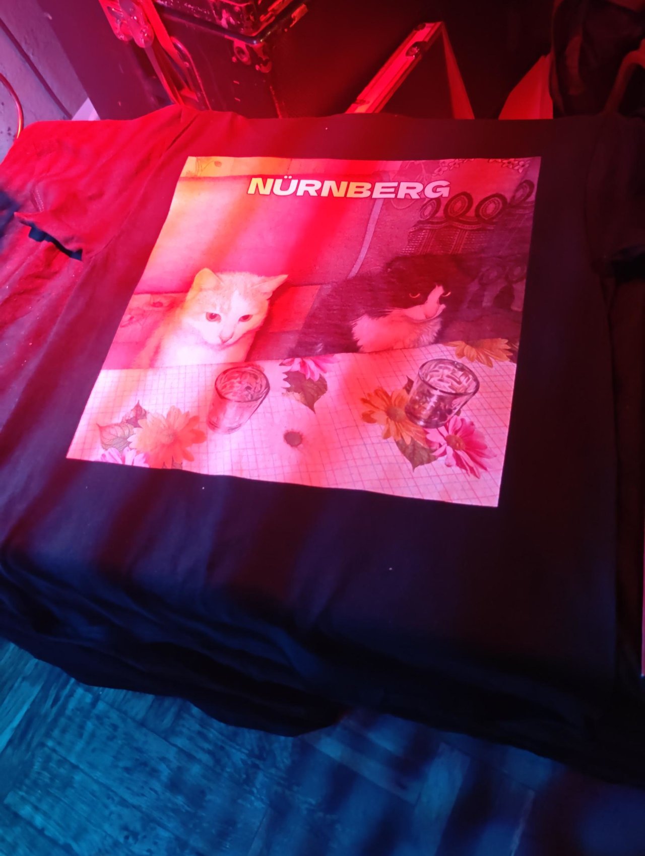 Printed T-shirt shold as merch. Image of two cats sitting at a table in front of two glasses of clear liquid. Typical eastern-european deco. Caption says "Nürnberg"